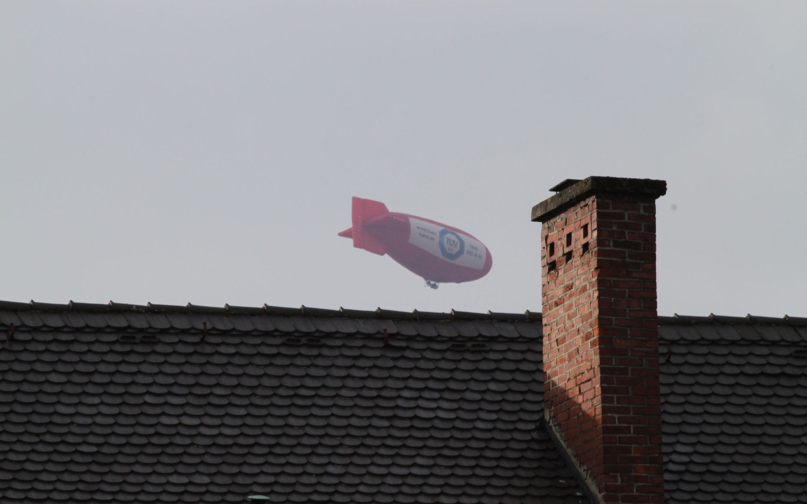A red advertising blimp floats above a tiled rooftop and brick chimney