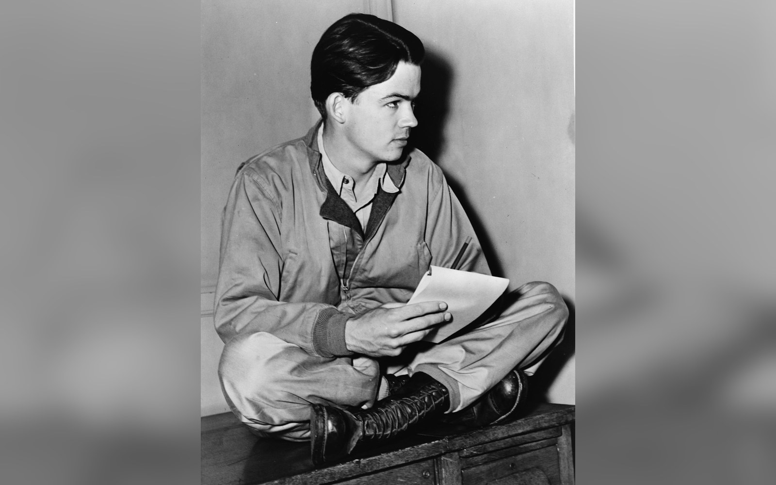 Young man in military coveralls sits cross-legged holding papers, looking to the side