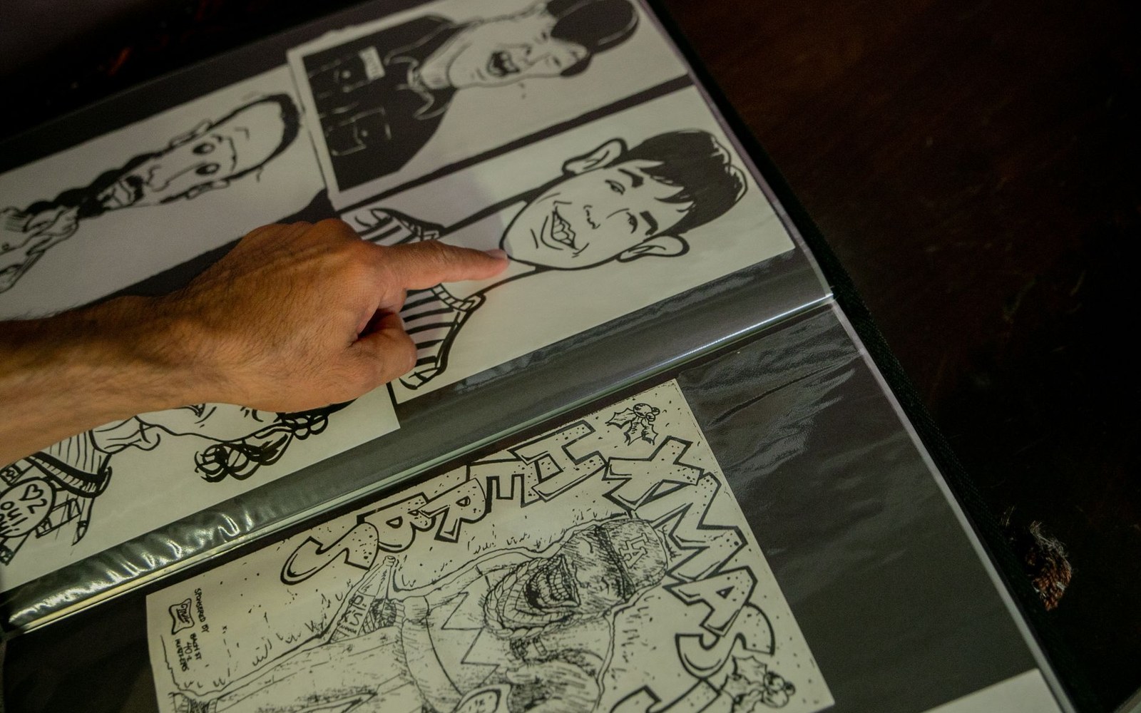 A finger points to a black-and-white comic strip panel in a binder of drawings