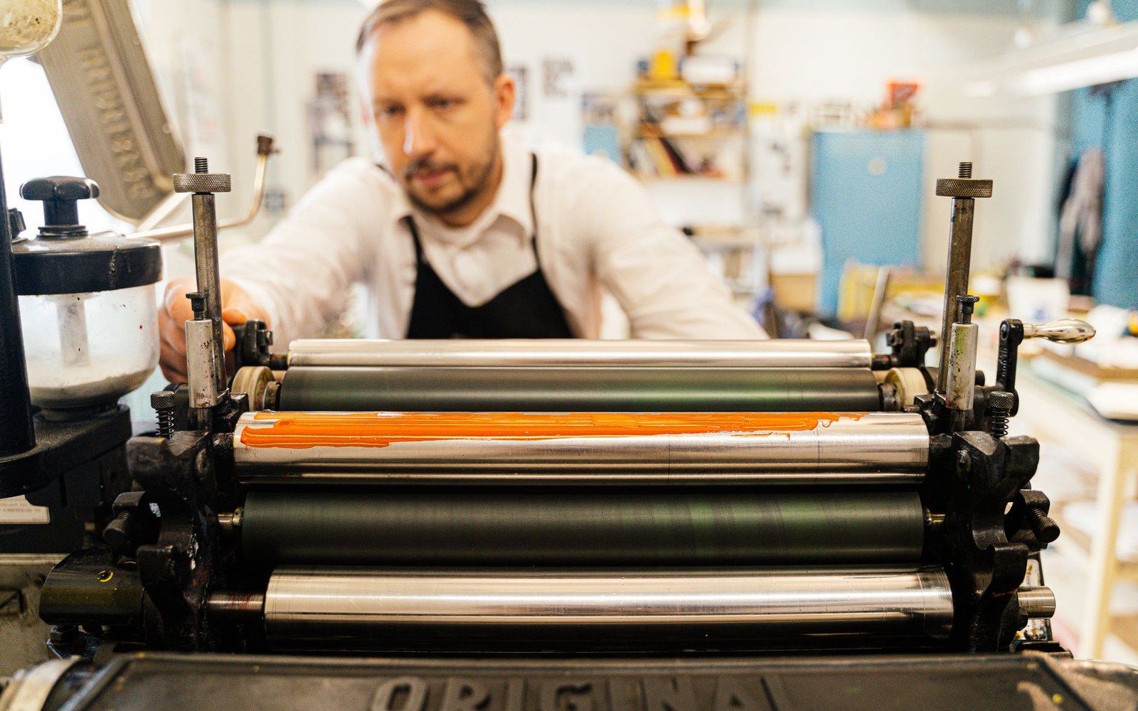 A letterpress operator at the press, adult, in work clothes, inked hands, the forme locked in the bed of a platen press — workshop interior