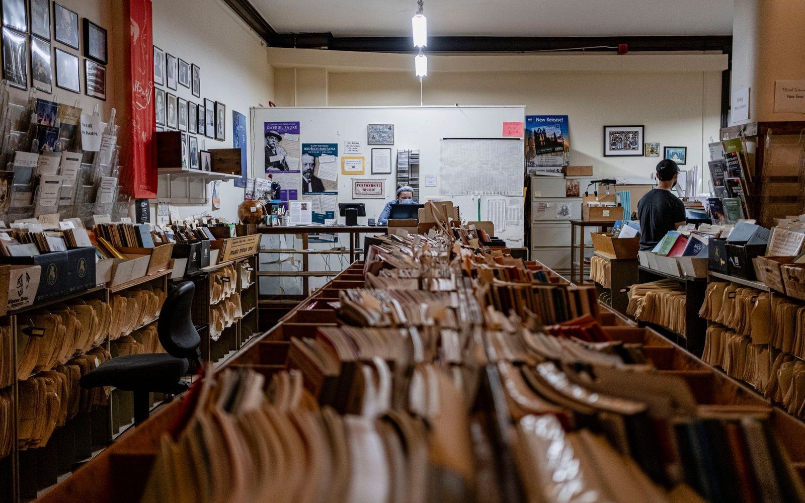 An archivist in a large archive interior, gloved, lifting a large unframed print from an open plan chest under cool overhead light — face visible